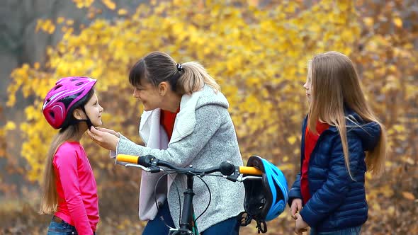 Mom helps two daughters put on a safe helmet before riding a bike on autumn day in nature