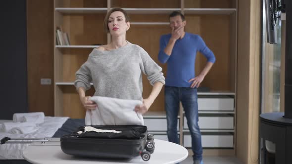 Portrait of Stressed Caucasian Attractive Woman Packing Travel Bag with Blurred Shocked Man Standing alt