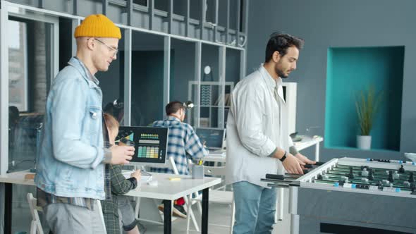Diverse Group of Employees Playing Foosball in Office While Colleagues are Working in Background alt