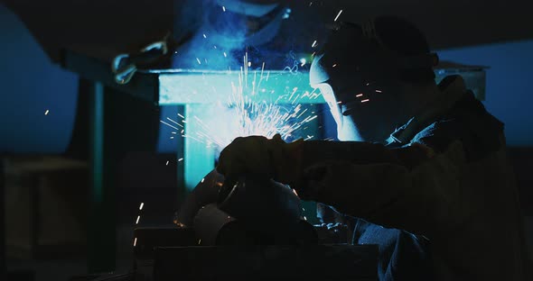 Welder in a welding helmet, in work clothes welds a pipe at the factory. Sparks alt