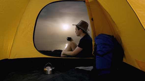 View from Tent of Young Man Hiker in Cowboy Hat Sits and Drinking Tea