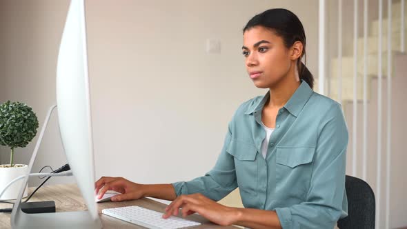 Motivated Young Woman Sitting at Desk in Home Office Using Computer for Web Surfing
