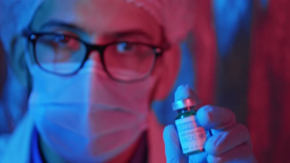Male doctor in a protective mask shows a bottle of vaccine and recommends it in emergency light alt