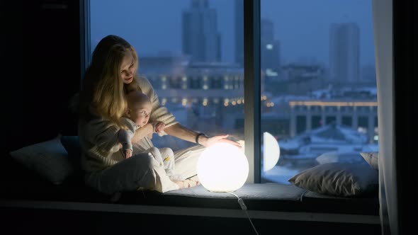 a woman with a child sit on the windowsill at night by a glowing lamp