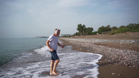 A Slender Whitehaired Elderly Man in White Headphones is Dancing Energetically Barefoot on the Beach