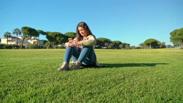 Side View of Young Girl Using Smartphone on a Grassy Field with the Sun Shining in the Background