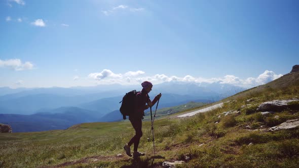 Young Woman with Backpack and Trekking Poles Walks Uphill Towards the Summit Against Blue Sky. Slow alt