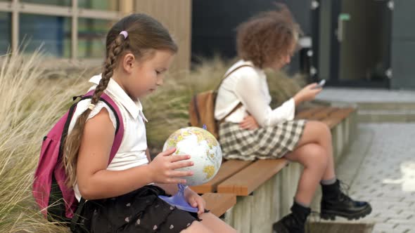 Two Schoolgirls of Different Ages are Sitting on a Bench in the School Yard alt