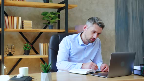 Focused man at home office in wireless earphones looks laptop computer writes information notebook alt