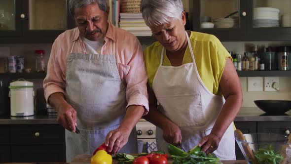 Happy senior mixed race couple wearing aprons preparing food in kitchen alt