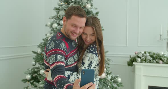 Young Couple Taking Selfie on Mobile Phone on Christmas Decor Background. Man Holds a Mobile Phone alt