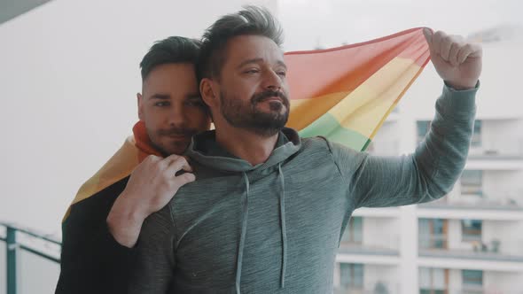 Proud Young Gay Male Couple Hugging on the Balcony Covered with Rainbow Flag alt