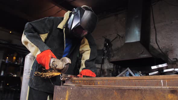 A Man in a Welder's Mask Uses a Grinder to Saw Off a Profile on a Desktop in the Workshop
