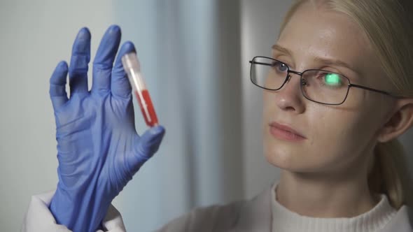 A Woman Scientist with Glasses Holds a Test Tube with Blood in Her Hand and Examines It Carefully