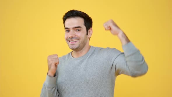 Handsome smiling Hispanic man raising his fists and dancing in yellow isolated studio background