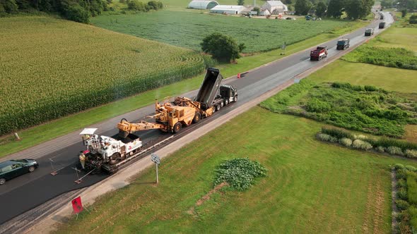 Road crew working on repaving section of rural highway. Drump trucks with supplies waiting.