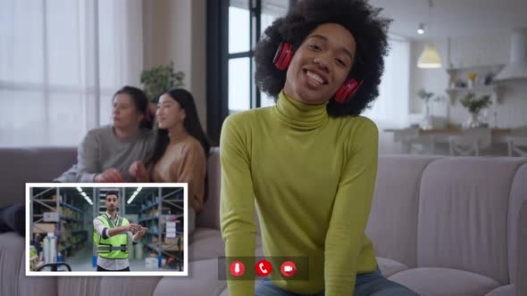 African American Woman Sitting in Home Office Talking Online with Middle Eastern Man Standing in alt
