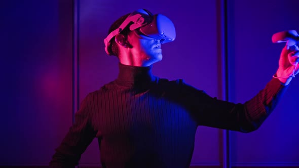 Young Man in VR Helmet Interacts with Virtual Reality Using Controllers alt
