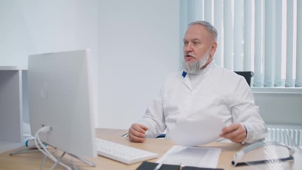 Grayhaired Male Doctor Sits at Workplace and Communicates with Colleagues Via Video Call Remote alt