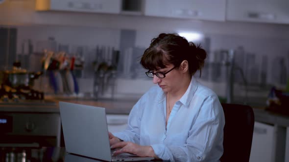 Woman with Glasses Sits at a Laptop in the Kitchen at Home