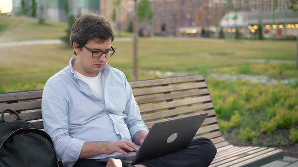 Young Man Typing in Laptop in Park Sitting on a Bench on Blurred Park Background