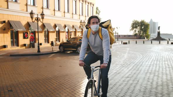 Portrait of a Courier with Yellow Backpack and Protective Mask Rides a Bicycle Through the City with alt