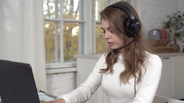A Young Woman in Headphones with a Headset Works at a Laptop alt