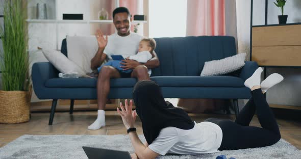 Woman in Hijab Lying on the Floor Using Laptop and Waving Hands to Her Handsome Positive alt