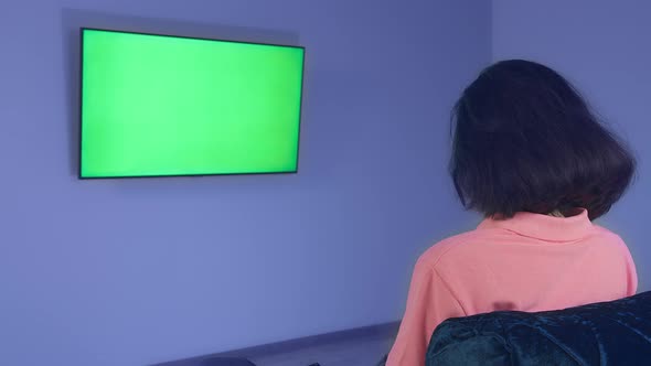 Girl Sitting in Front of a TV Hanging on the Wall Watching News Chromakey Rear View alt
