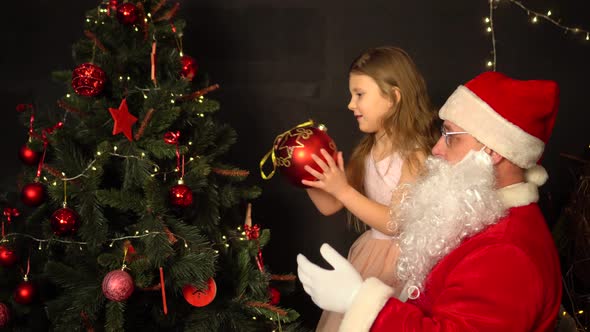 Santa Claus and a little girl decorate a Christmas tree. Santa holds the girl in his arms