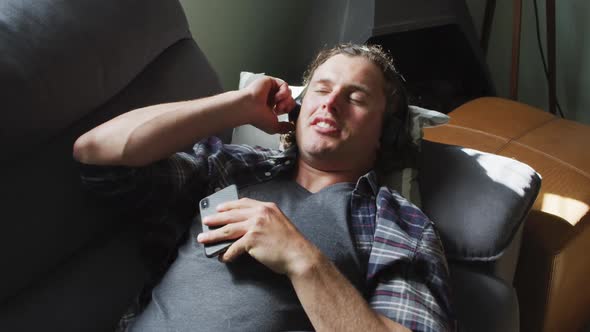 Relaxed caucasian man using smartphone and wearing headphones lying on sofa in living room alt