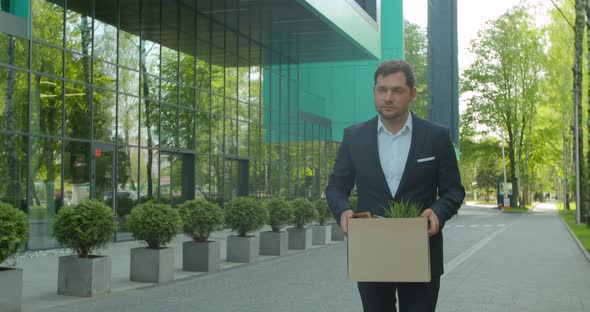 Emotional Disappointed Corporate Worker Gets Fired and Walking Near Office Center with Carton Box alt