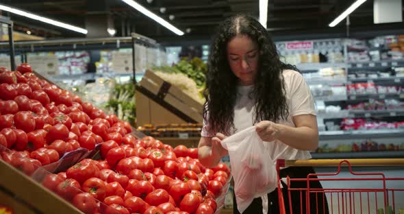 Young Woman in the Grocery Section of a Supermarket Picks Up a Bag and Puts Tomatoes in a Basket to alt