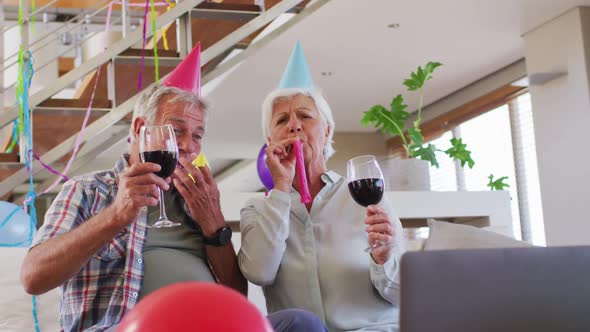 Senior caucasian couple blowing party blowers celebrating birthday toasting wine glasses at home alt