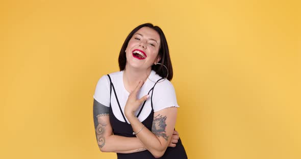 A young embarrassed woman with tattoo is listening compliments shy and laughing in yellow studio alt
