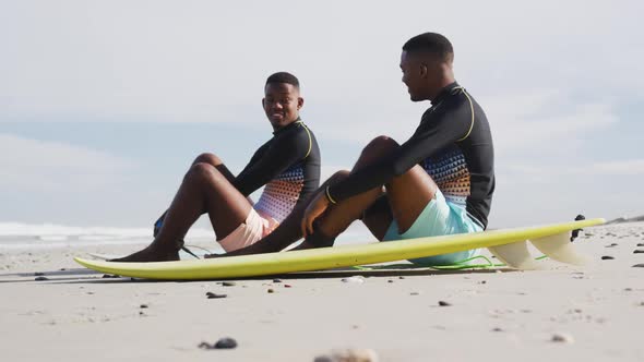 Happy african american teenage twin brothers sitting by surfboards on a beach talking alt
