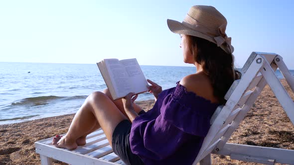 A Girl in a Straw Hat Sits on a White Sun Lounger By the Sea with a Book in Her Hands
