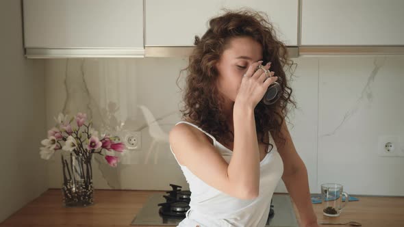 A Young Woman is Standing in the Kitchen and Drinking Coffee alt