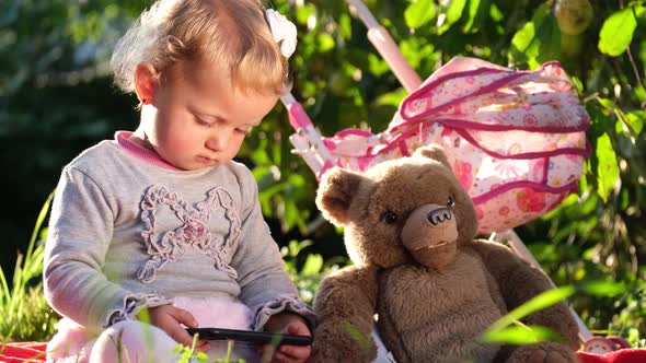 Little Girl Looks at Her Phone She is Sitting on a Blanket in the Garden alt