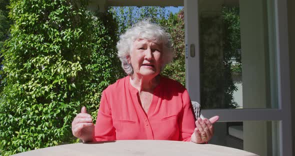 Caucasian senior woman sitting at table in sunny garden talking and gesturing alt