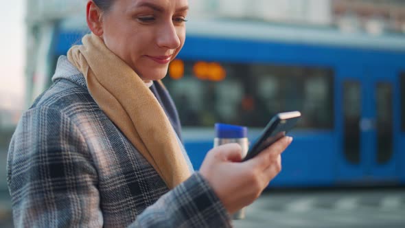 Woman in a Coat with a Thermos in Her Hand Uses a Smartphone alt