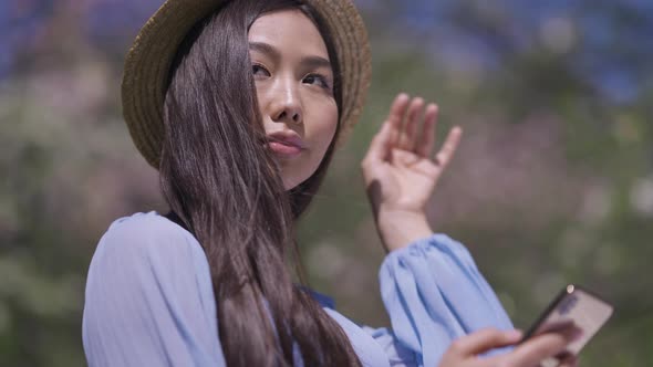 Young Asian Woman in Straw Hat Texting on Smartphone Smiling Standing in Sunlight in Spring Sakura alt