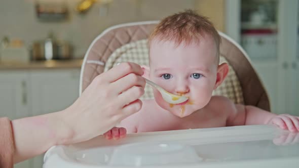 Mother feeding toddler baby from a spoon on a high chair for children, home kitchen background.