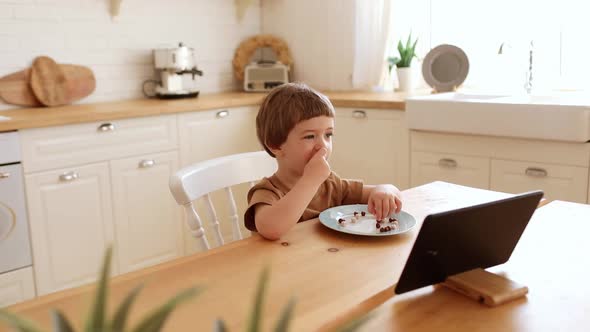a Little Caucasian Boy Eats Krunchy and Watches Tablet PC at Kitchen Table alt