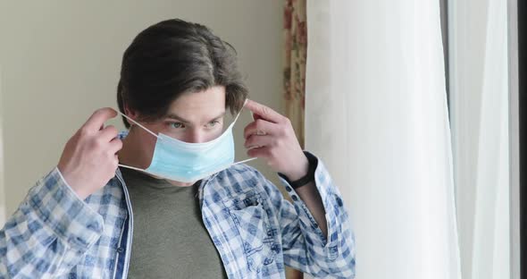 Young Man on Self-isolation at Home Wears Medical Mask and Looks Out a Window alt