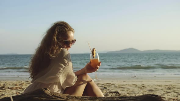 Woman Sitting in White Dress Drinks Pineapple Cocktail Pina Colada on Beach alt