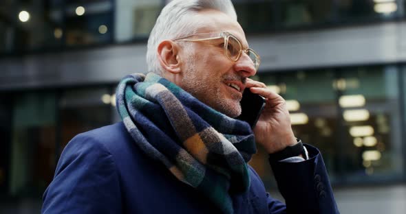 A Grayhaired Man Uses a Mobile Phone Standing in Business Center of the City alt