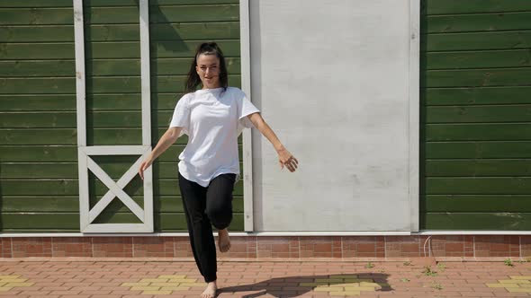 Barefoot Brunette in Black Pants and a Big White Tshirt Dances Against a Green Wall on the Street on alt