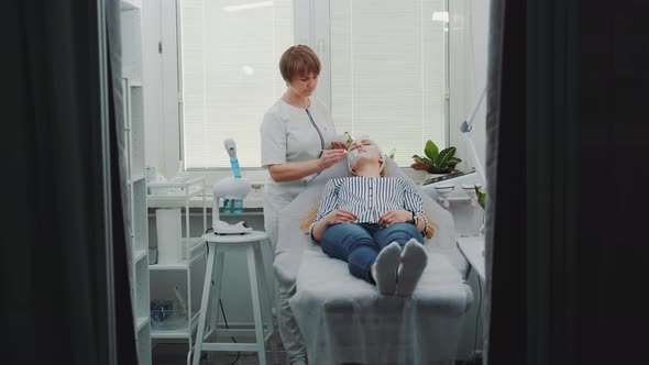 Cosmetologist Applying Cream Mask on Face of Pretty Woman at Beauty Spa Salon alt