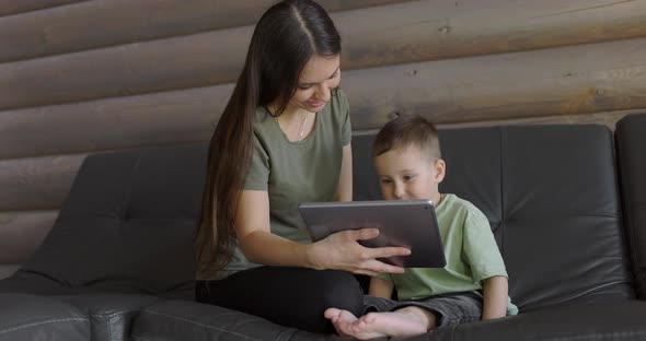 Young Mum and Her Son Play Online Games on Digital Tablet While Sitting on Sofa in Living Room alt